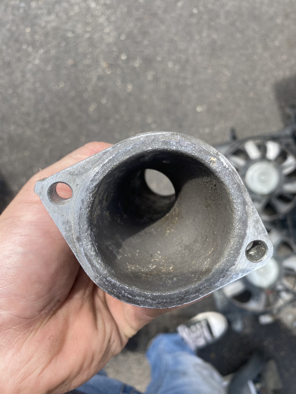 Hand holding a worn thermostat housing, radiator fans on the ground behind